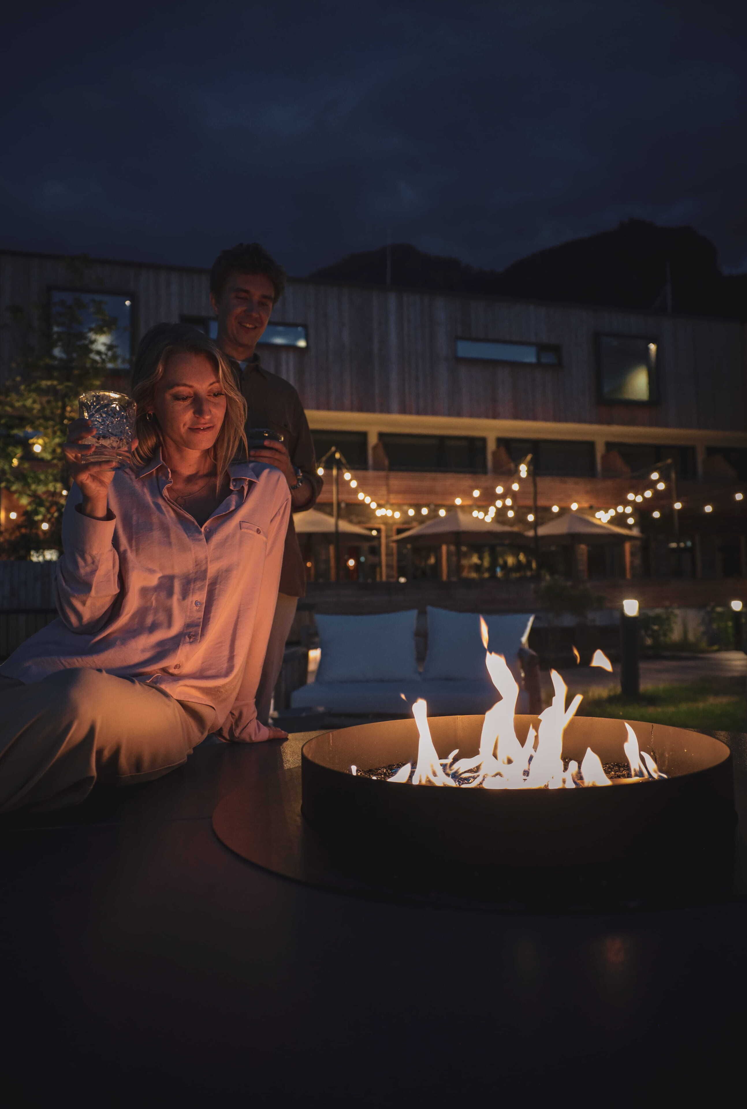 A couple sitting by a fire pit with a building and string lights in the background.