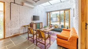 Een ruime kamer met een bakstenen muur, een houten eettafel met stoelen, een bank en een aan de muur bevestigde televisie.