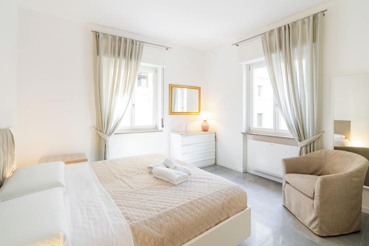 A hotel room with a bed, a chair, a nightstand with a lamp, and a window with curtains.