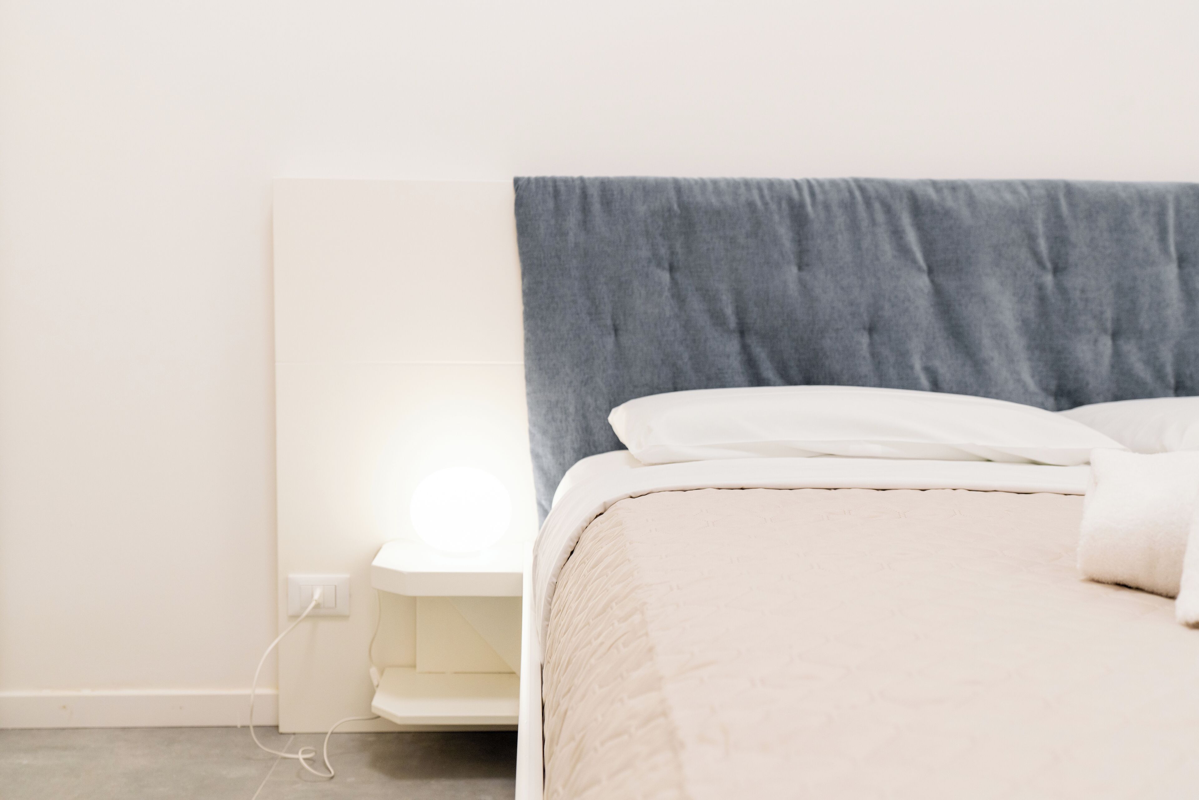 Een net opgemaakt bed met een grijs hoofdbord en witte beddengoed, een witte nachtkastje en een stopcontact aan de muur.