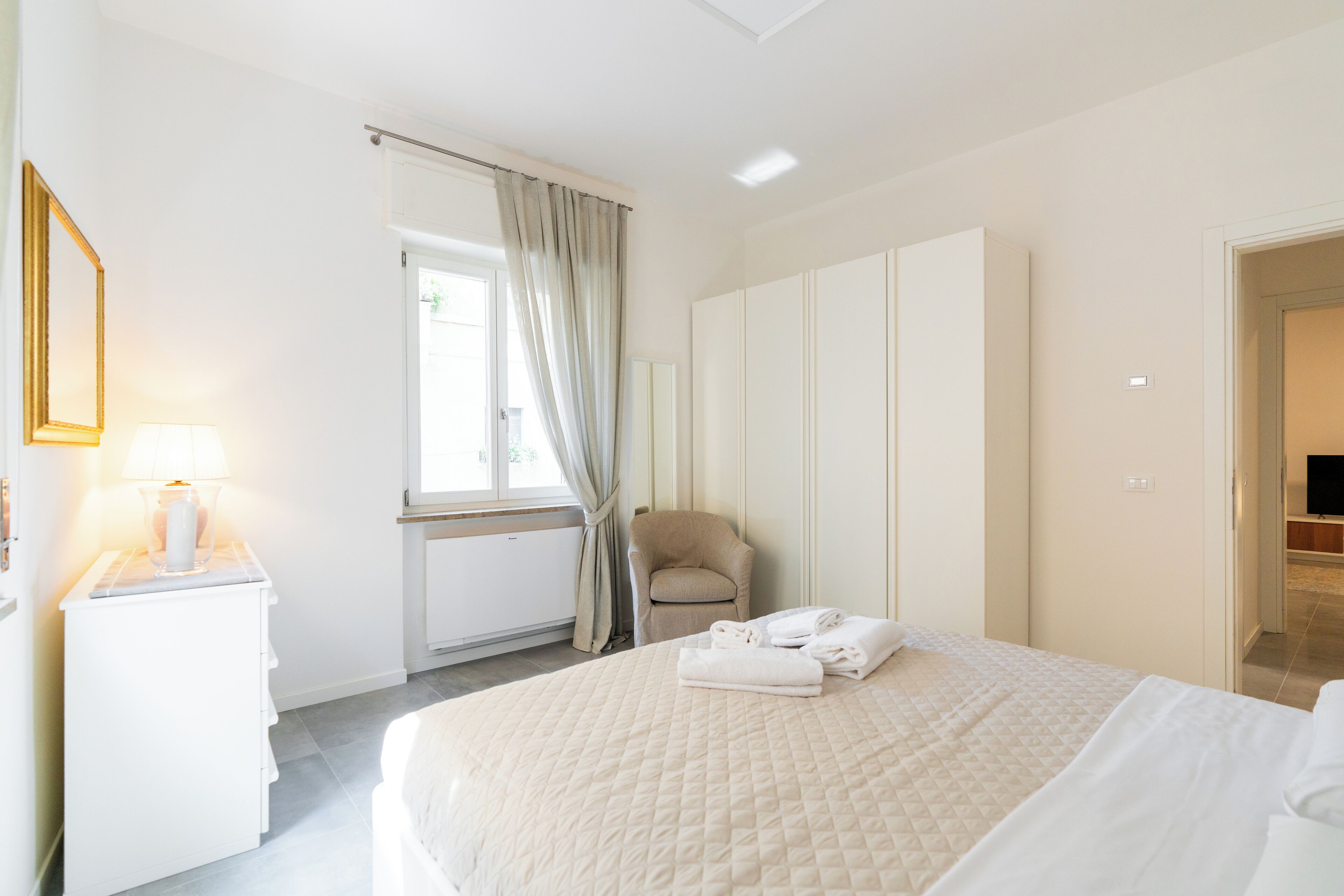 A hotel room with a bed, a chair, a desk with a lamp, a wardrobe, and a television in the corner.