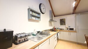 Una cocina con encimera de madera, pileta de acero inoxidable y una tostadora. Hay un reloj en la pared y una alacena con vajilla.