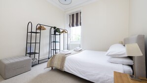 A neatly arranged hotel room with a bed, a bedside table, a lamp, a radiator, a window with striped curtains, a grey bench, and a metal shelf with clothes and a plant.