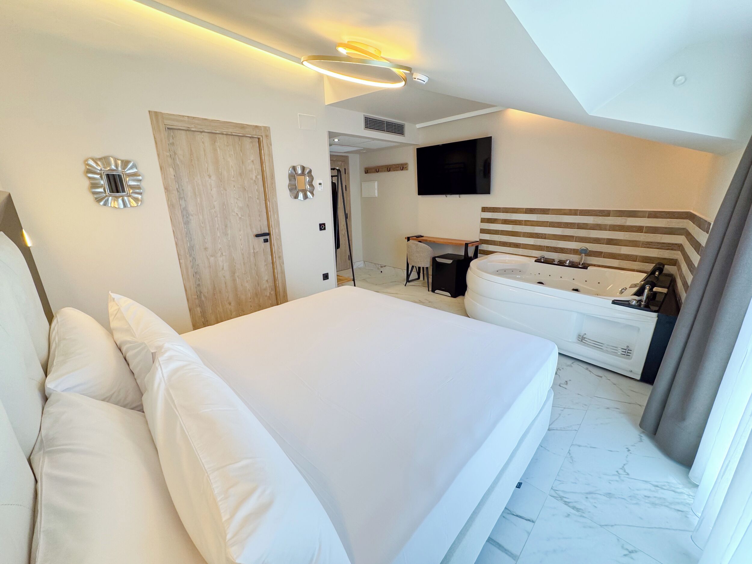 A modern hotel room with a large bed, a sitting area with a chair, a desk, and a large bathtub.