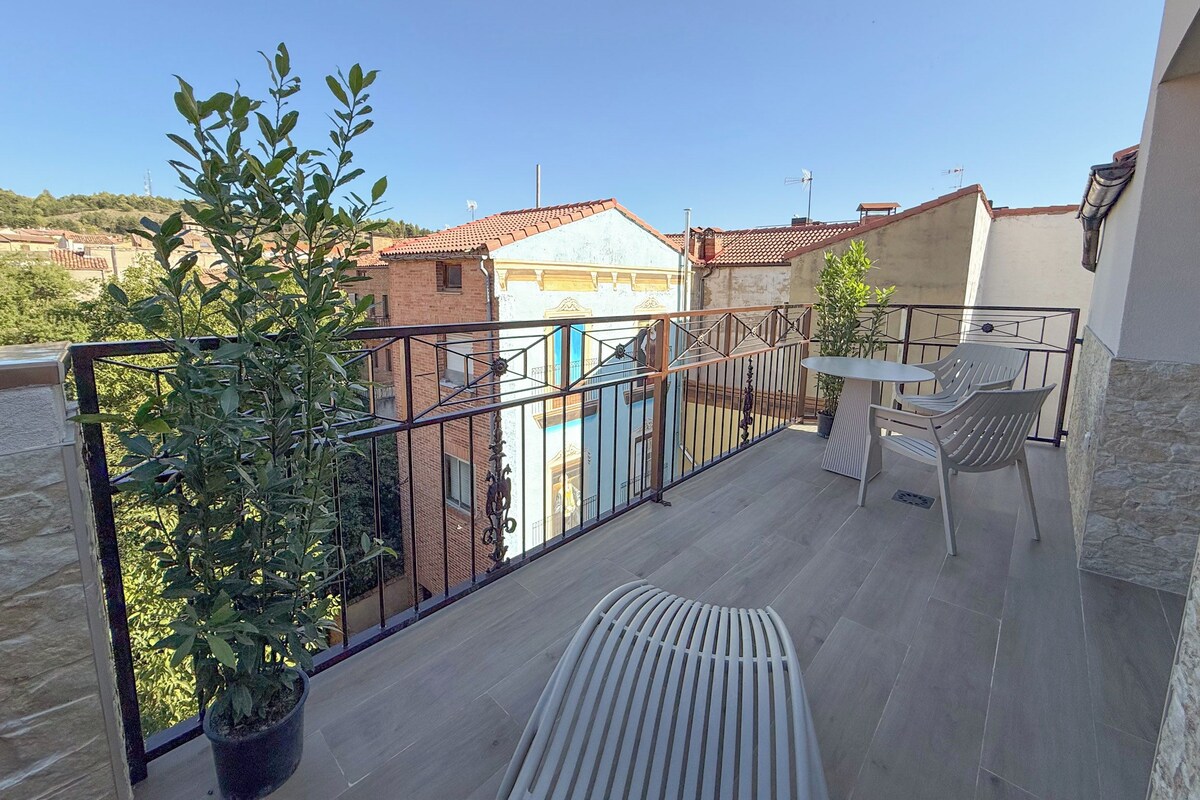 Un balcón con una mesa, sillas, plantas en macetas y vistas a edificios cercanos.