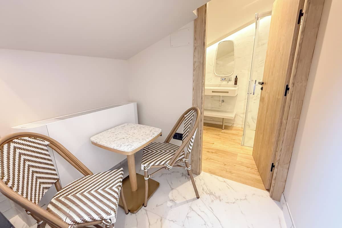 Una habitación pequeña y moderna, con una pared blanca, una mesa pequeña, dos sillas y una puerta de madera que da a un baño.