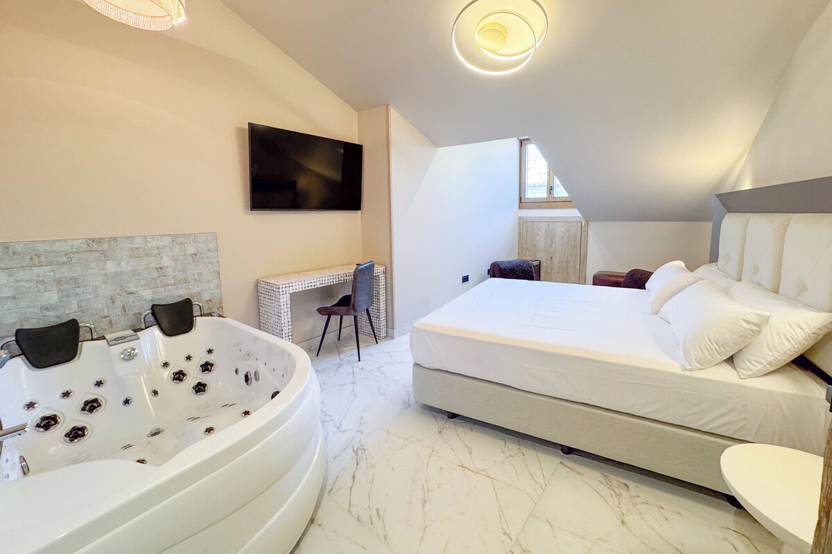 A modern bedroom with a large bed, a sitting area with a desk, and a bathtub.