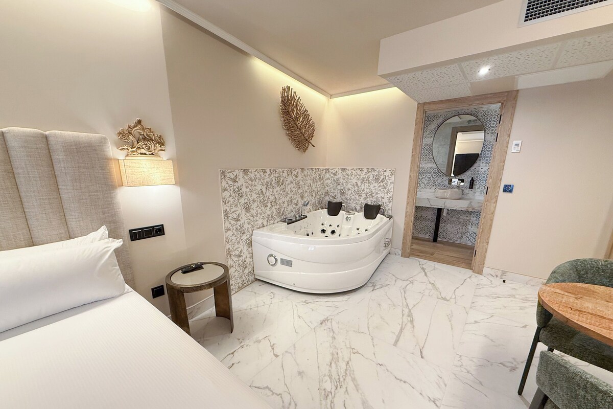 A modern hotel room with a large bed, a sitting area with a small table, a jacuzzi, and a bathroom with a mirror and sink.