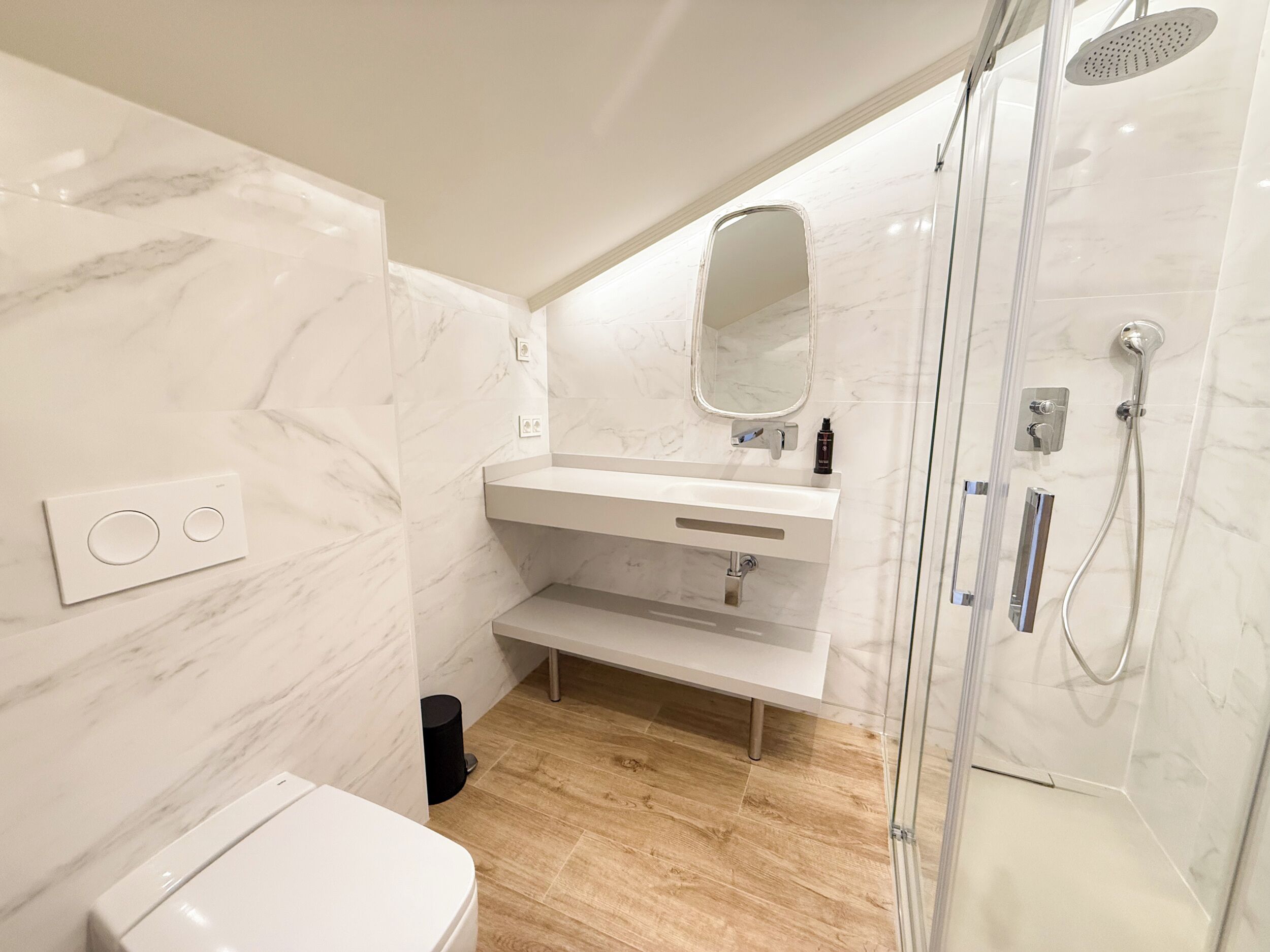 A modern bathroom with a marble wall, a white sink, a mirror, a shower, and a toilet.