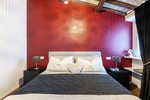 Un dormitorio con pared roja, cabecera blanca y colcha acolchada negra. Hay dos mesitas de noche con lámparas y un estante pequeño con objetos decorativos.