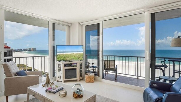 A spacious living area with a large TV, a sofa, and a dining table. The room has a balcony with a view of the beach and ocean. - Beachfront 2BR/2BA condo with pool, Wi-Fi, and ocean views. (Marco Island)