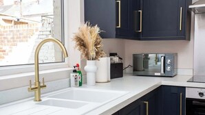 Una cocina con encimera blanca, grifo dorado, armarios oscuros con tiradores dorados, un jarrón blanco con flores secas y un microondas.