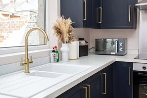 A kitchen with a white countertop, a gold faucet, dark cabinets with gold handles, a white vase with dried flowers, and a microwave.
