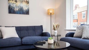 Un salón moderno con un sofá azul oscuro, una mesa redonda de centro, una lámpara de pie y vistas a un edificio a través de un ventanal amplio.