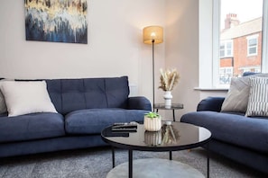 A modern living room with a dark blue sofa, a round coffee table, a floor lamp, and a view of a building through a large window.