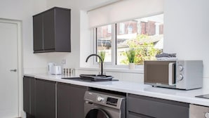 Een kleine keuken met een wasmachine, een magnetron en een raam met uitzicht op een tuin.