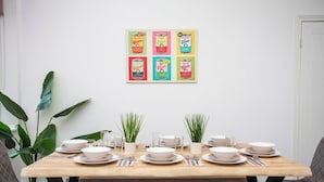 Een eettafel gedekt met borden, kommen en bestek, omringd door potplanten, met een kleurrijk kunstwerk op canvas met blikthema erboven.