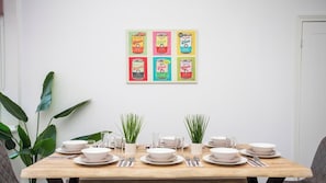 Une table Ă manger dressĂ©e avec des assiettes, des bols et des couverts, agrĂ©mentĂ©e de plantes en pot, et surmontĂ©e d'une Ćuvre d'art colorĂ©e dont le thĂšme est celui des boĂźtes de conserve.