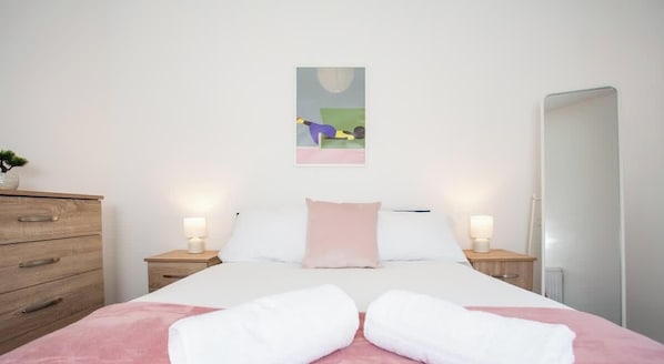 A bedroom with a bed, two bedside tables, a nightstand, a mirror, and a wall-mounted artwork.