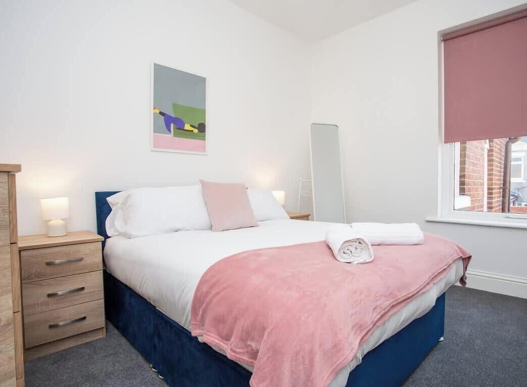 A bedroom with a bed, bedside table, lamp, artwork, window, and a view of the outside.