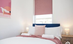 A bedroom with a bed, bedside tables, a window with a blind, and a wall-mounted artwork.