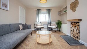 A living room with a grey sofa, two armchairs, a round coffee table, a hanging light fixture, a fireplace with logs, and a window with curtains. - Vacant Nest | The South End | Spacious | Smart TV (Kielder)