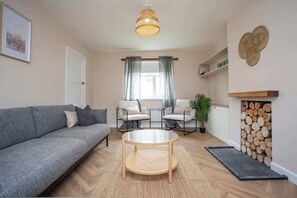 A living room with a grey sofa, two armchairs, a round coffee table, a hanging light fixture, a fireplace with logs, and a window with curtains. - Vacant Nest | The South End | Spacious | Smart TV (Kielder)