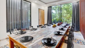 A dining area with a long table set for a meal, large windows with curtains, and a view of greenery outside. - Alaya Stays Fortezza | Pet-friendly 6-BHK Farmhouse With Private Pool (Sohna)