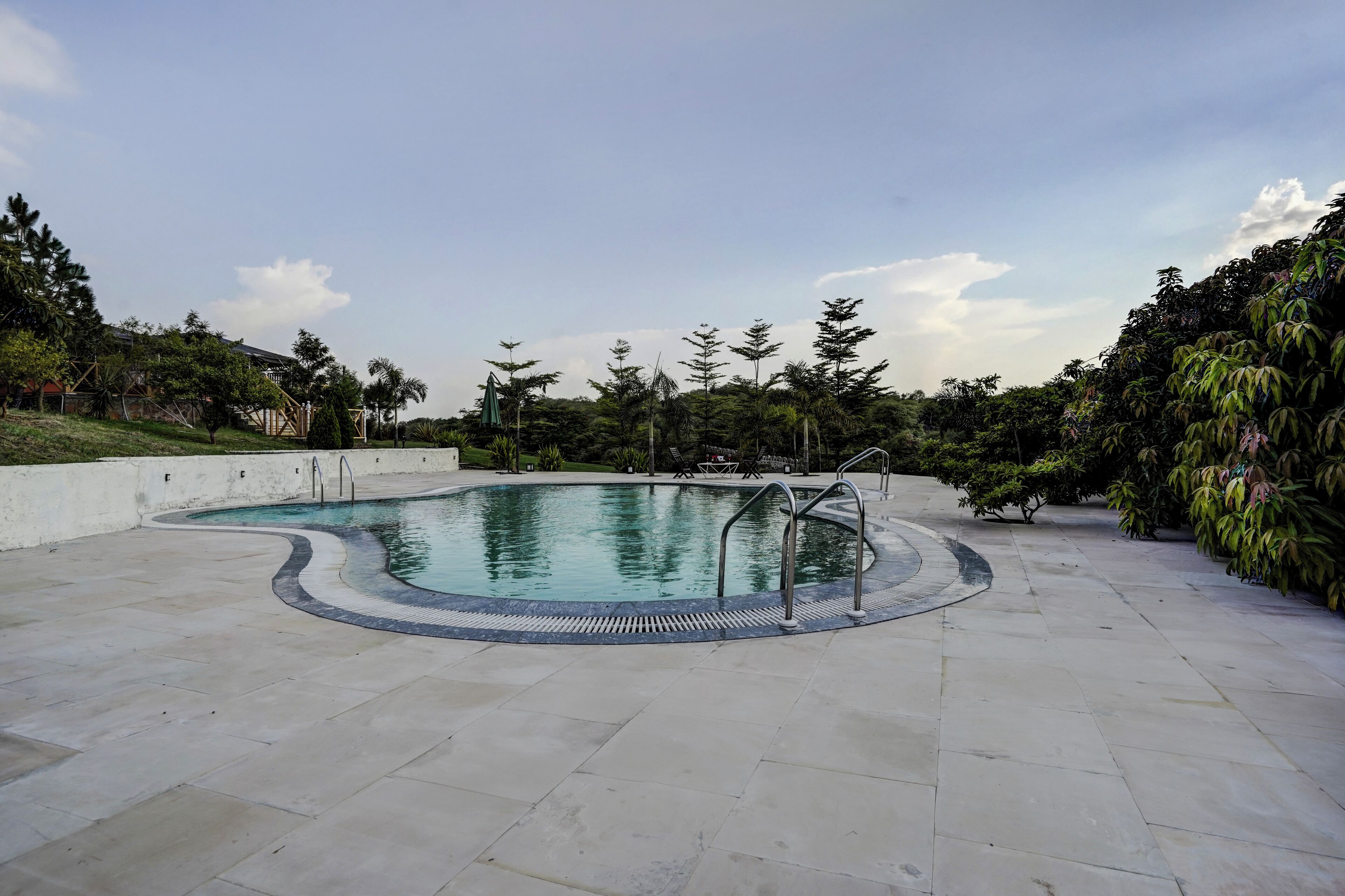 A swimming pool with a tiled deck, surrounded by greenery and a clear sky.