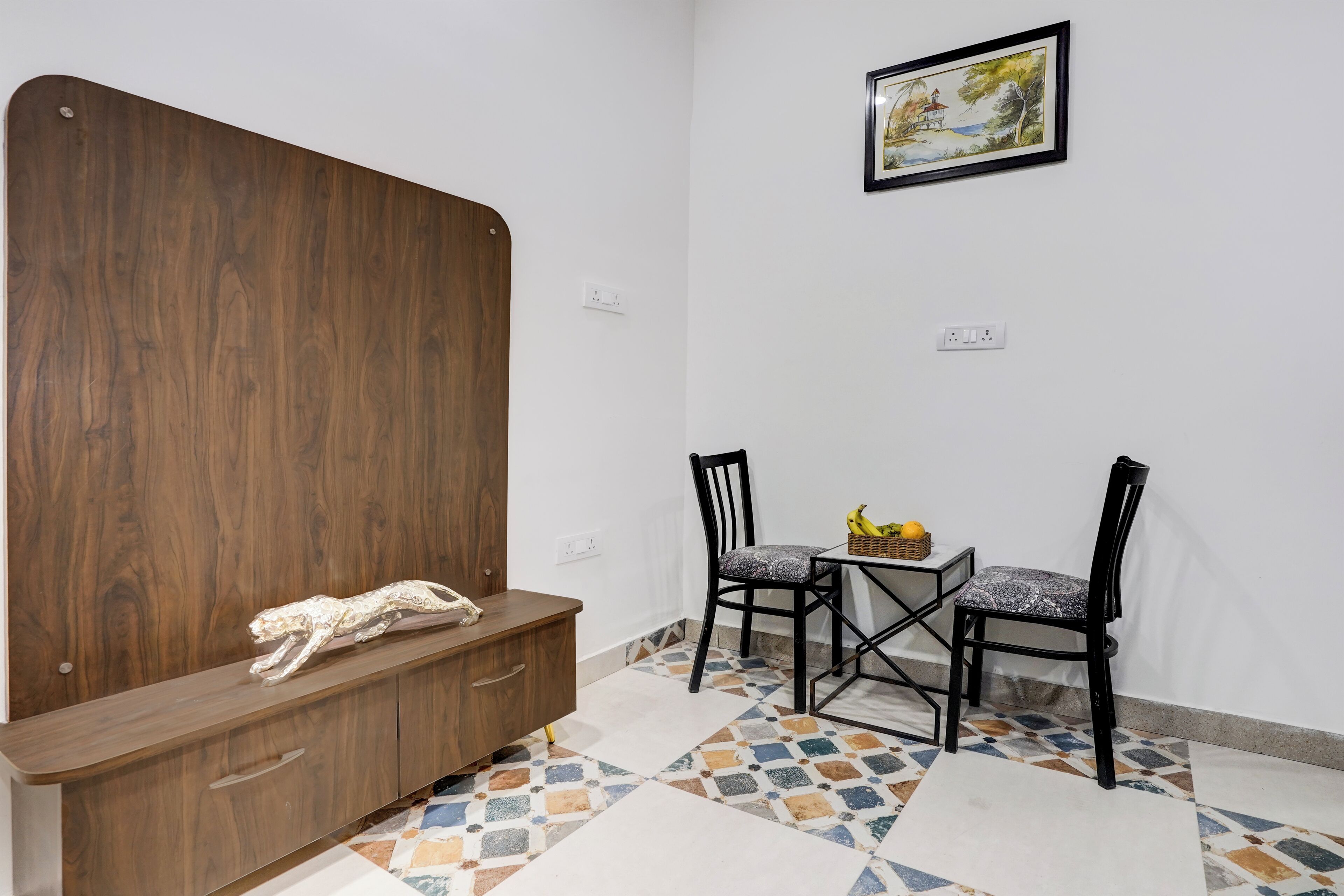 A room with a wooden bench, two chairs, a small table with fruits, and a painting on the wall.