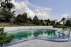 A swimming pool with a curved edge, surrounded by a tiled deck and a low wall. - Alaya Stays Fortezza | Pet-friendly 6-BHK Farmhouse With Private Pool (Sohna)