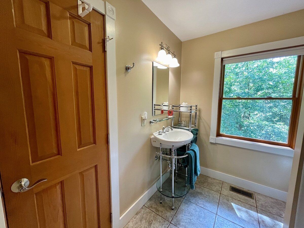 A bathroom with a wooden door, a white sink, a mirror, and a window with a view of trees.