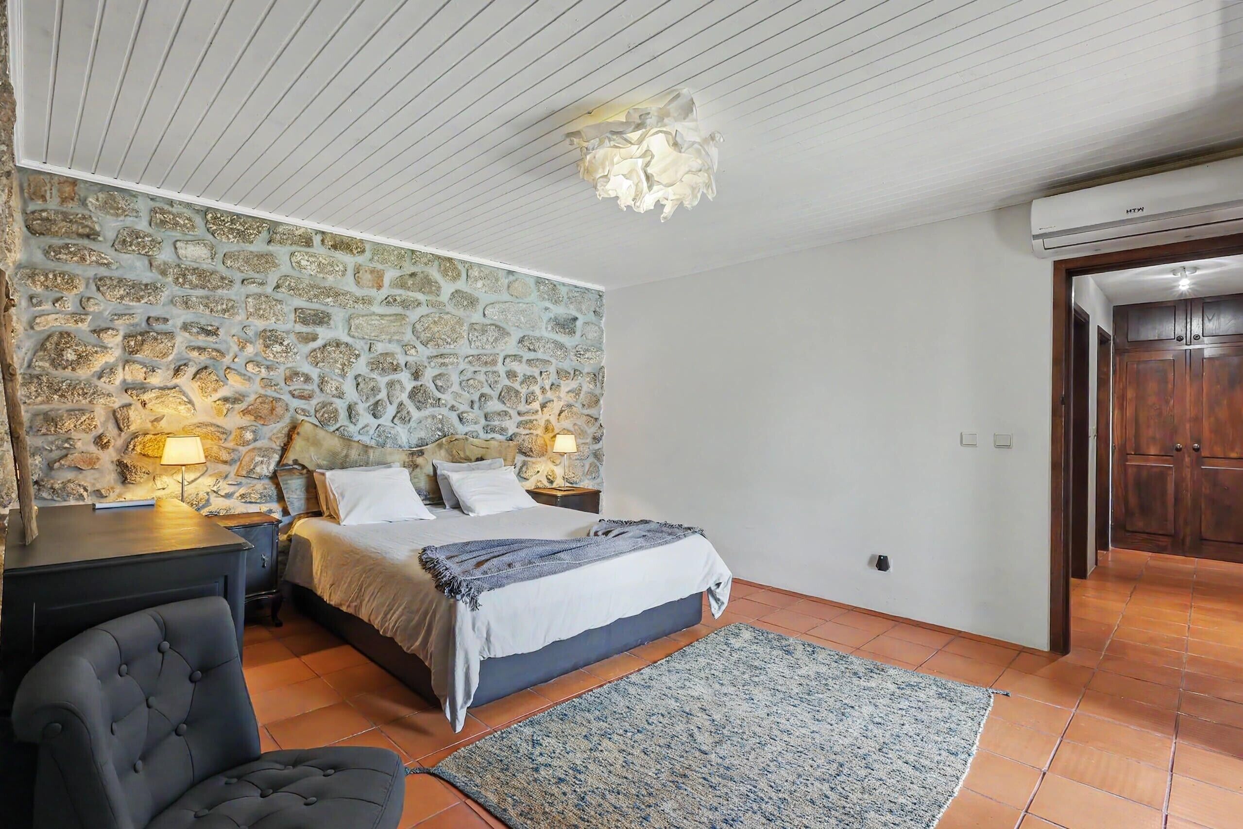 Camera da letto con parete in pietra, un letto con testiera, un comodino, una sedia e un tappeto grande.