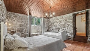 Un dormitorio con paredes de piedra, techo de madera, cama con ropa de cama blanca, mesita de noche y una ventana con cortinas.