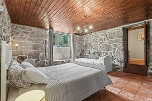 Une chambre avec des murs en pierre, un plafond en bois, un lit avec une literie blanche, une table de chevet et une fenêtre avec des rideaux.