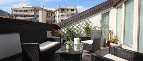 A rooftop terrace with wicker furniture, potted plants, and a view of buildings in the background.