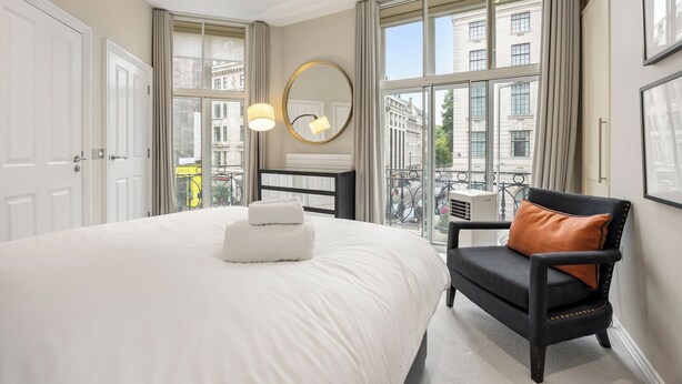 Habitación de hotel con una cama grande, un espejo redondo, un sillón negro con cojín marrón y ventanas amplias con vistas a edificios.