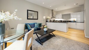 Una sala de estar moderna con una mesa de centro de vidrio, un sofá con cojines azules, una zona de cocina con armarios blancos y una zona de comedor con mesa y sillas de vidrio.