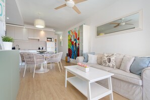 Une cuisine moderne avec des meubles blancs, un coin repas avec des chaises en osier, un canapé blanc agrémenté de coussins à motifs et un ventilateur de plafond.