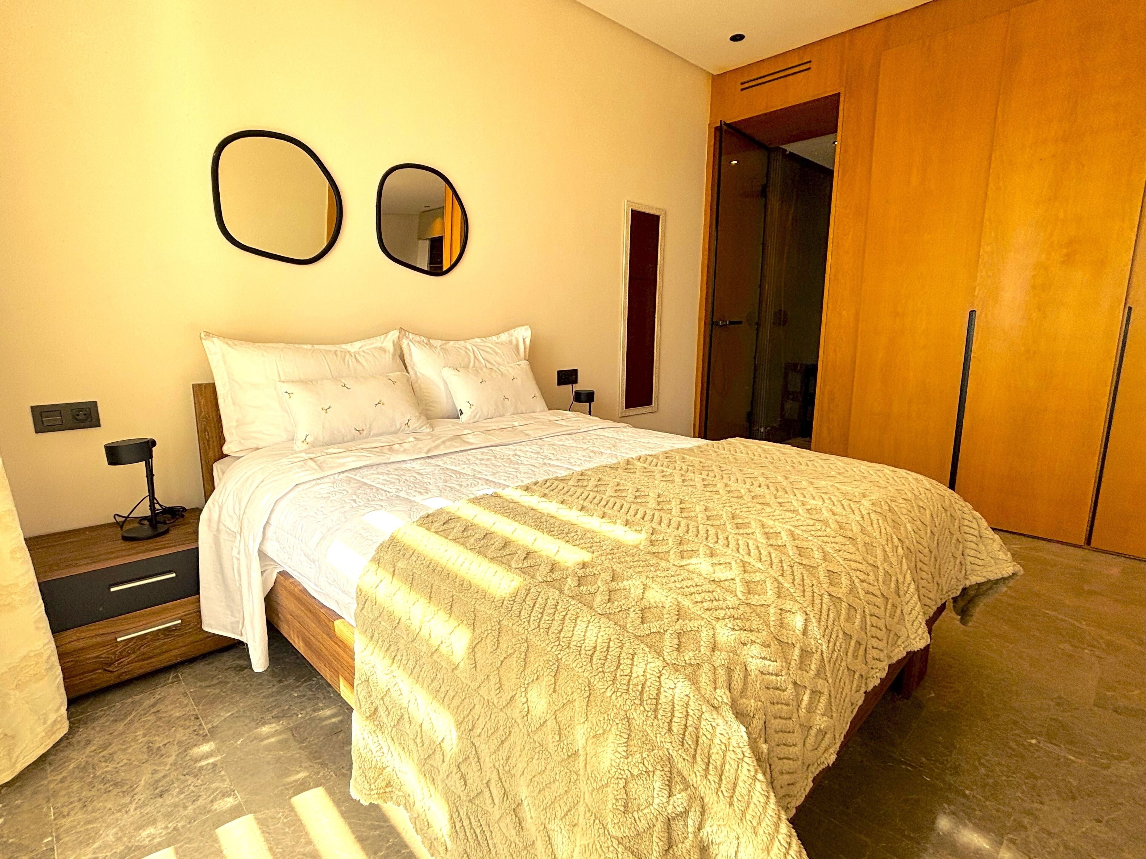 A modern bedroom with a large bed, a wooden wardrobe, a mirror, and a nightstand with a lamp.