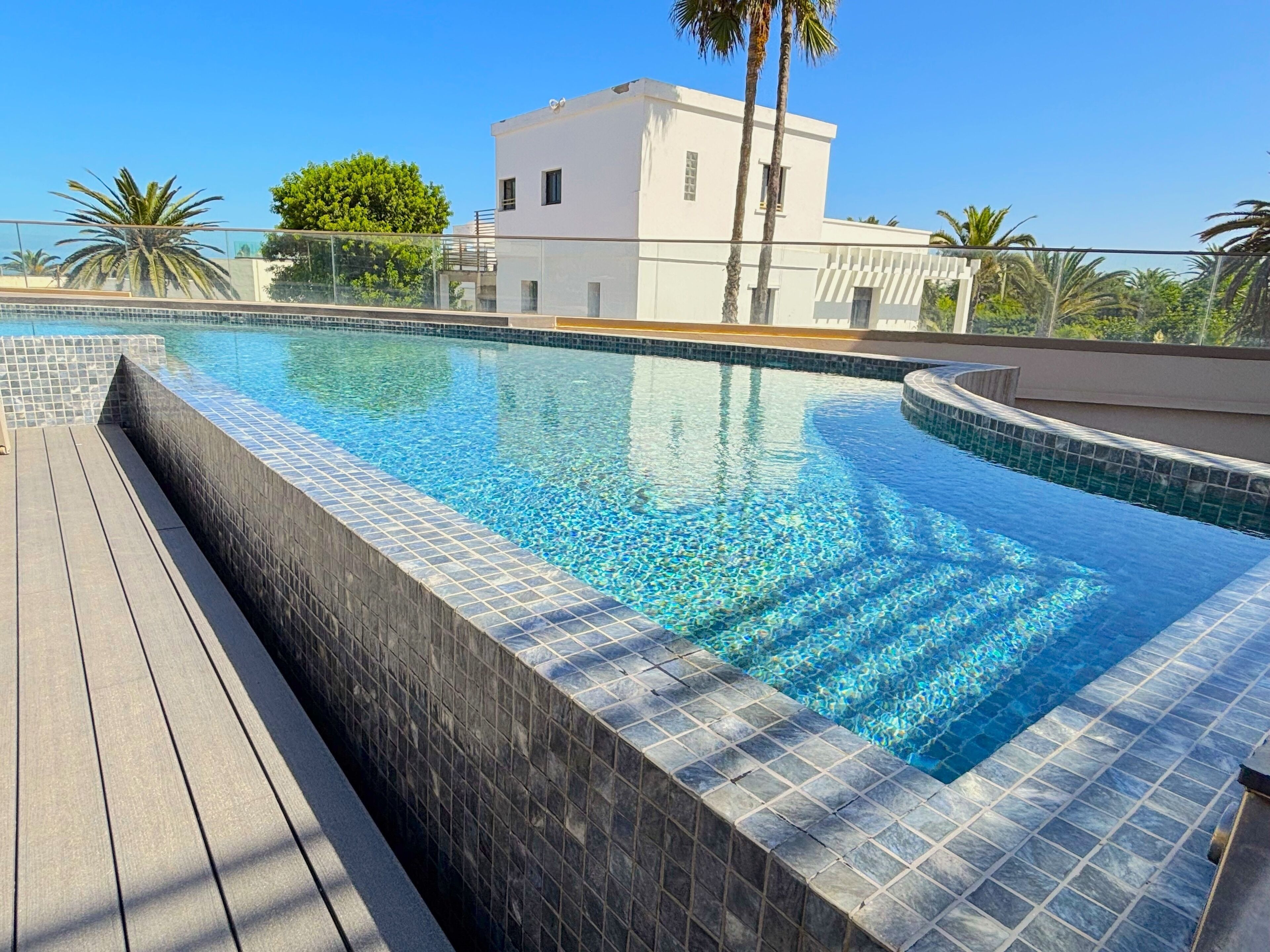 A swimming pool with tiled edges, a wooden deck, and a white building in the background.