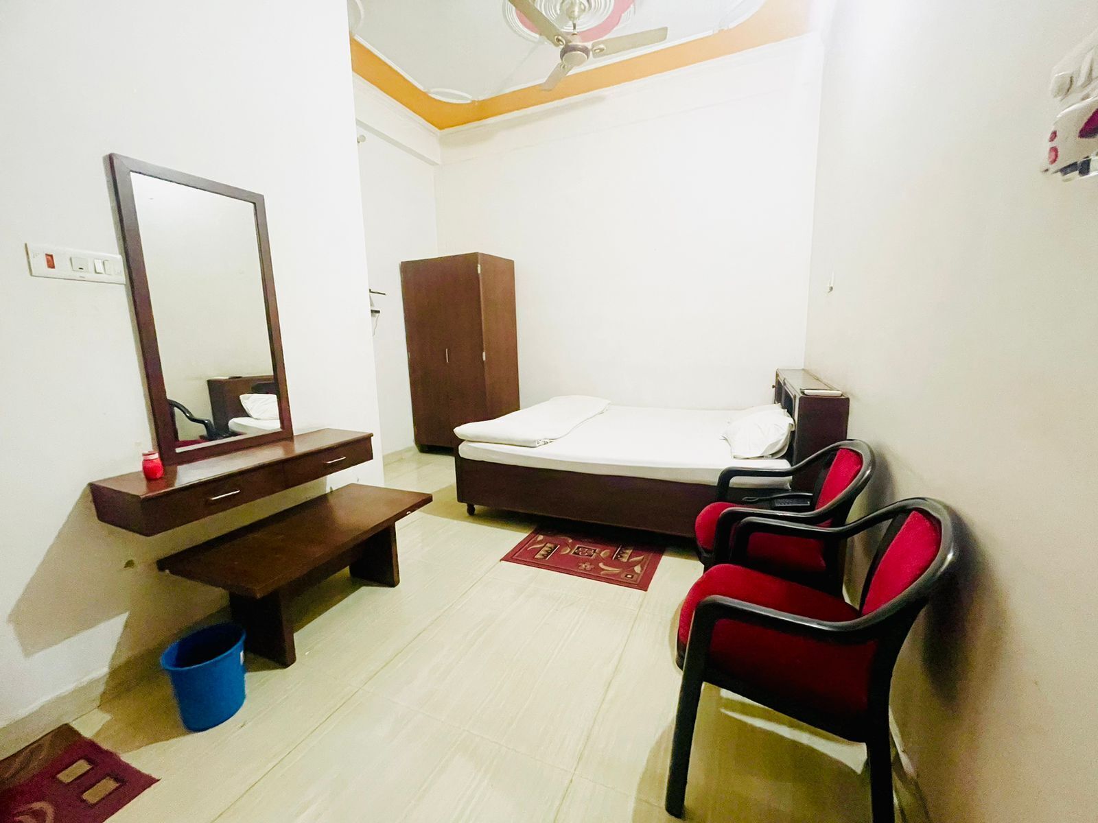 A small, simple room with a bed, a mirror, a wooden cabinet, a desk, a chair, and a blue bucket.