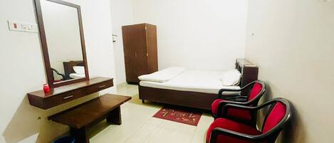 A small, simple room with a bed, a mirror, a wooden cabinet, a desk, a chair, and a blue bucket.