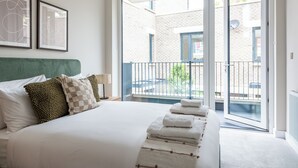 Une chambre d’hôtel avec un grand lit, une tête de lit verte, un balcon donnant sur un immeuble en briques et deux œuvres d’art encadrées accrochées au mur.