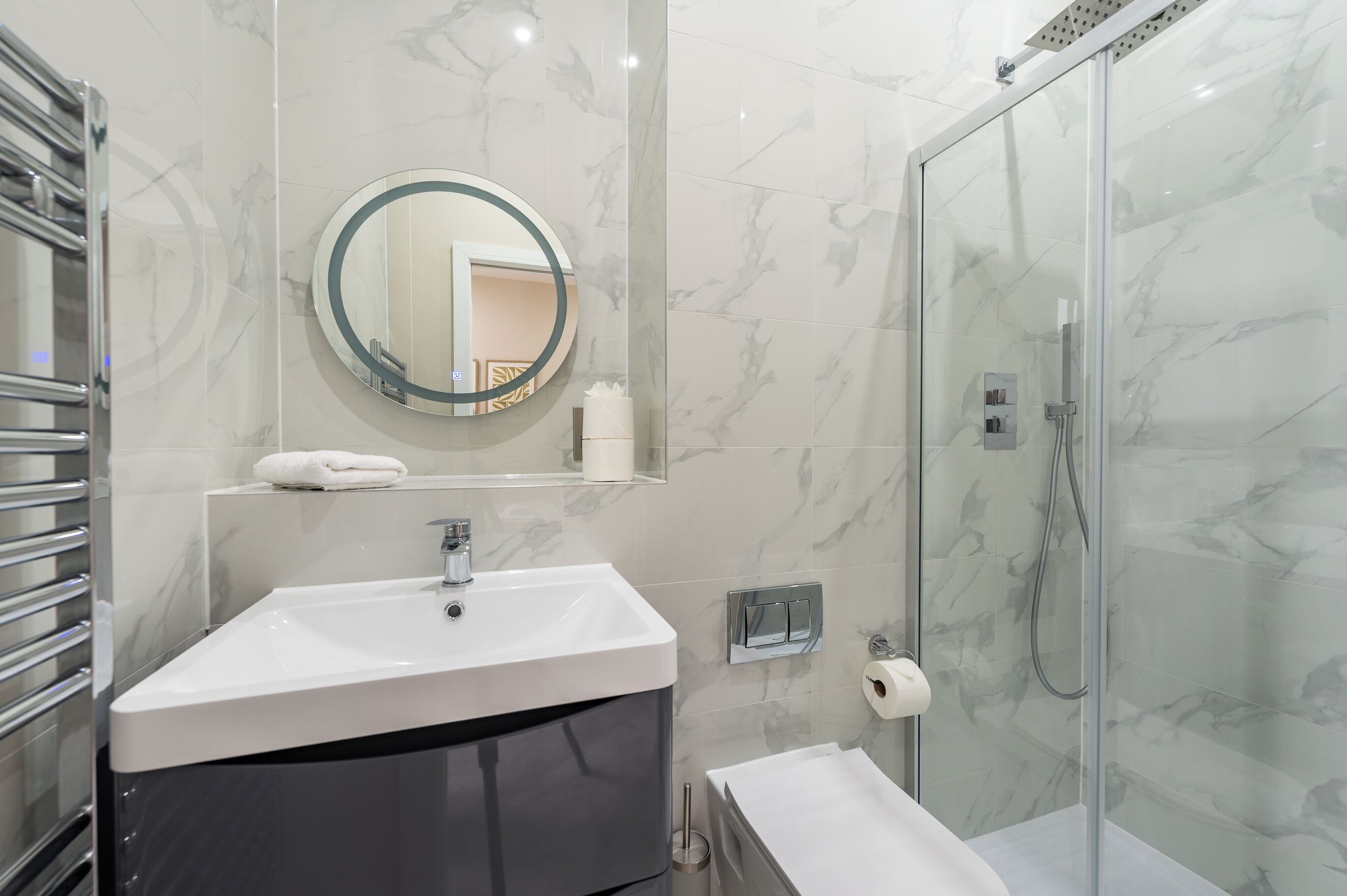 A modern bathroom with a marble sink, a glass-enclosed shower, and a round mirror.