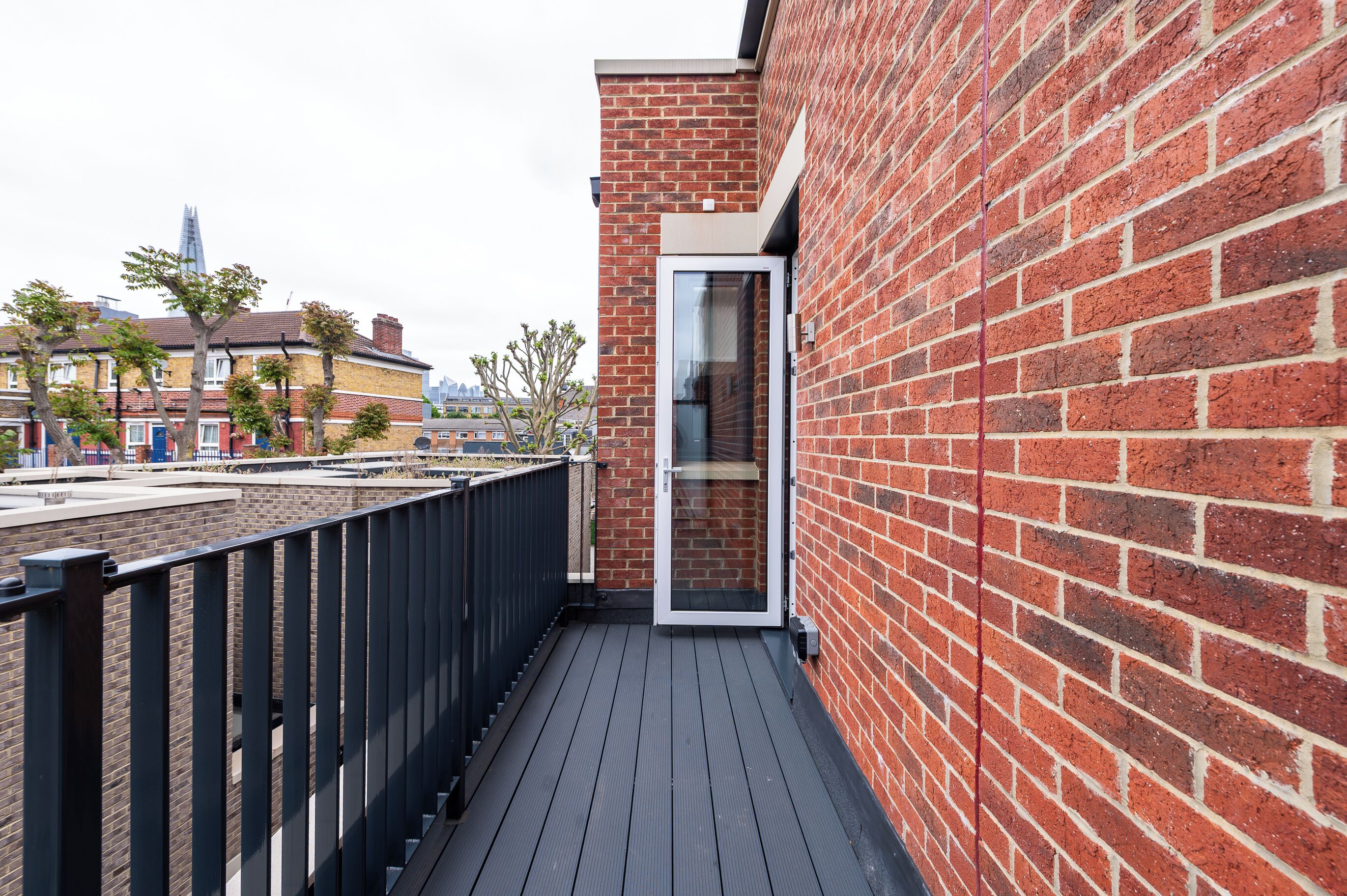 A narrow balcony with a brick wall and a glass door, overlooking a cityscape with buildings and trees.