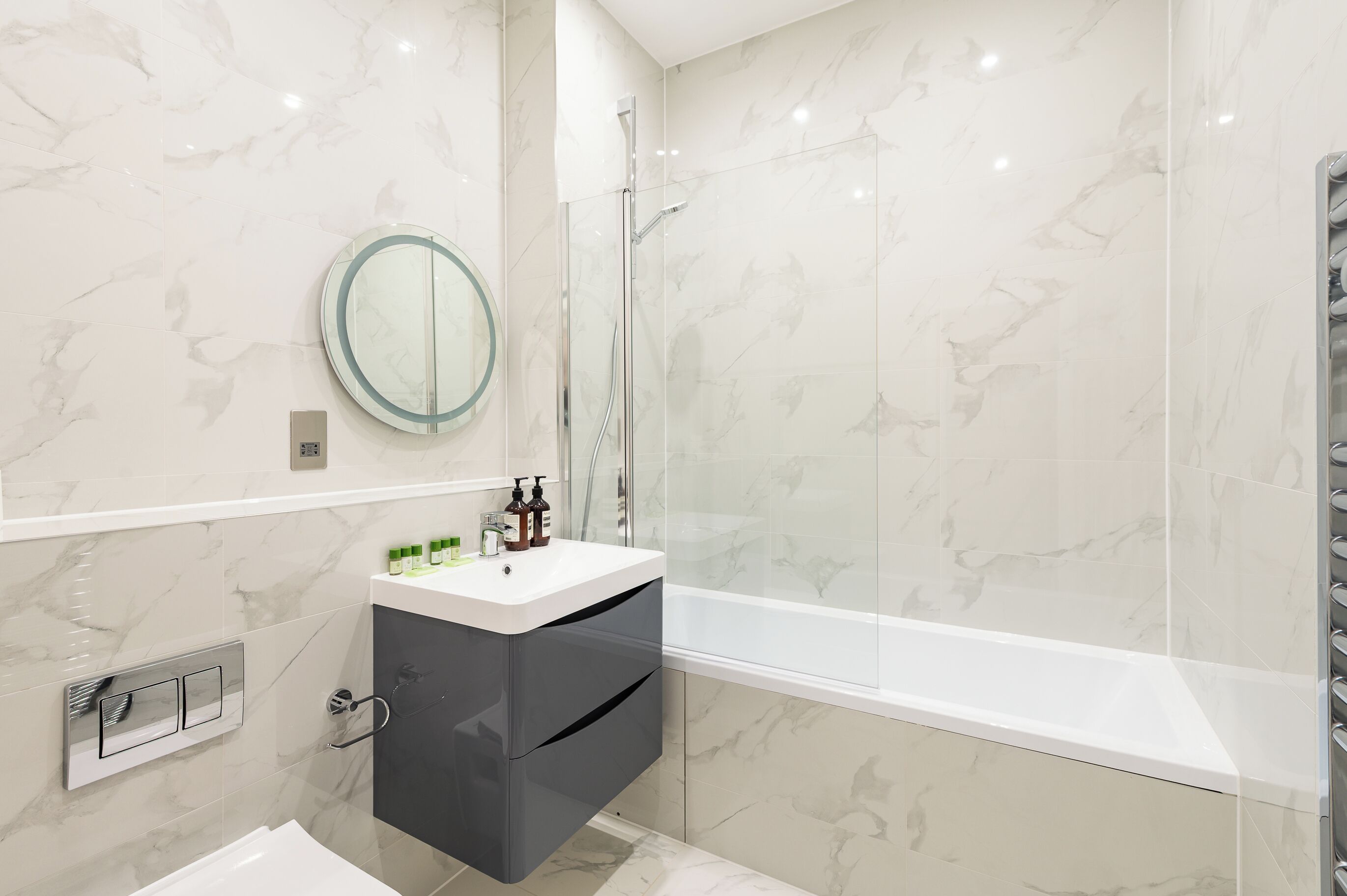 A modern bathroom with a marble backsplash, a round mirror, a white sink, and a bathtub.