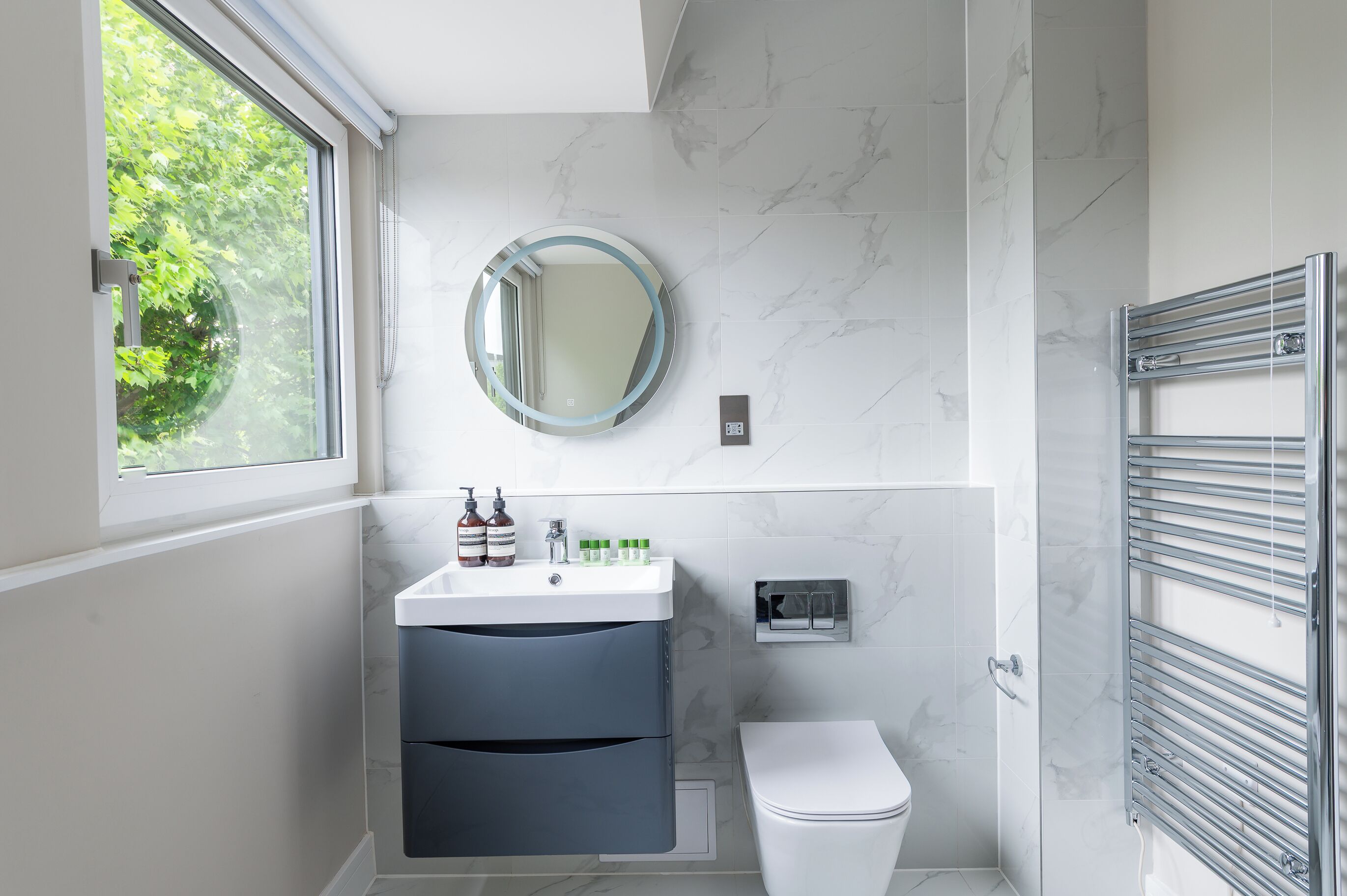 A modern bathroom with a white toilet, a sink with toiletries, a mirror, and a window with a view of greenery.
