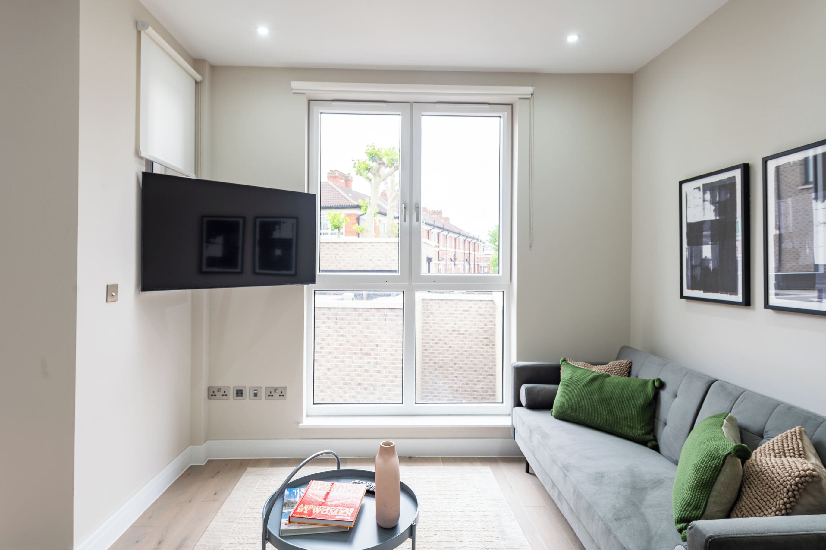 Un soggiorno moderno con un divano grigio, un tavolino con un vaso e libri, un'ampia finestra con vista sugli edifici e una TV a schermo piatto montata al muro.