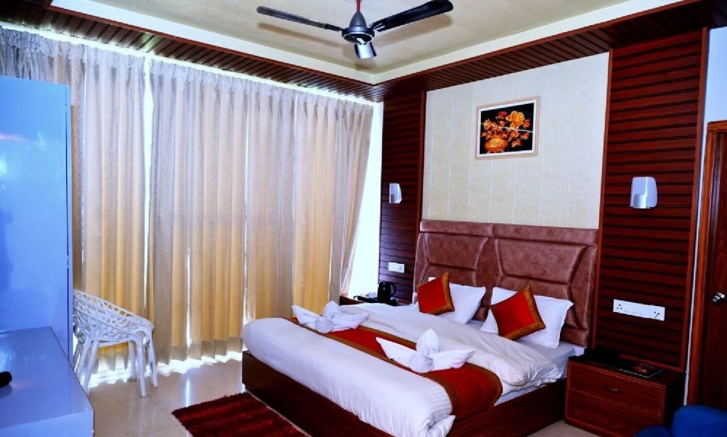 A hotel room with a large bed, a ceiling fan, a wooden headboard, a bedside table, a nightstand, a chair, and a window with curtains.